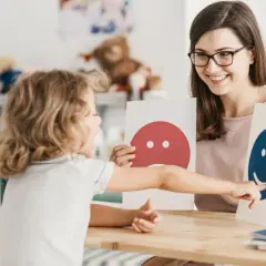 Dziecko wskazuje na uśmiechniętą buźkę, podczas gdy terapeutka trzyma kartę ze smutną. Widać tu ambiwalencję emocji.