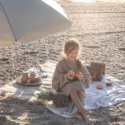 Dziewczynka na plaży pod parasolem, z owocami i kocykiem. Idealne rzeczy, co zabrać na wakacje nad morze z dzieckiem.