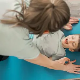 Chłopiec z dyspraksją śmieje się, leżąc na macie, podczas gdy dorosły pomaga mu w ćwiczeniach.