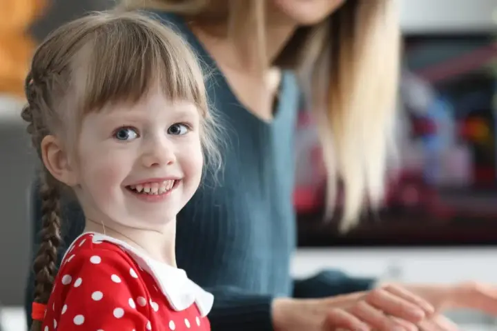 Uśmiechnięta dziewczynka w czerwonej sukience w groszki, z warkoczykami. Uczy się kindersztuby, co to znaczy, od mamy.