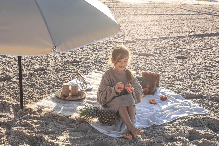 Dziewczynka na plaży pod parasolem, z owocami i kocykiem. Idealne rzeczy, co zabrać na wakacje nad morze z dzieckiem.