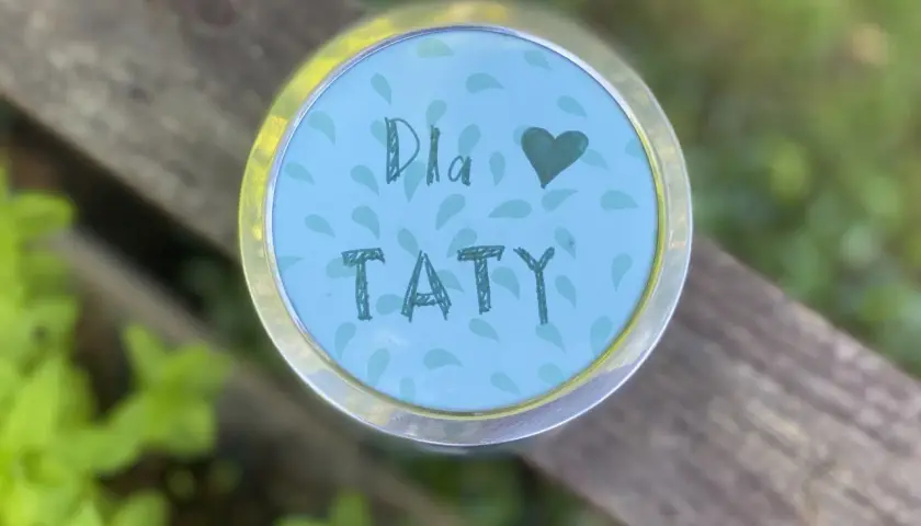 Uroczy upominek na Dzień Taty z przedszkola. Na okrągłej, błękitnej nakrętce z listkami, ręcznie napisane "Dla Taty" i serduszko.