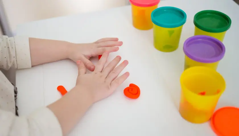 Dziecko bawi się kolorową ciastoliną dla dzieci, tworząc z niej małe kształty. Wokół leżą pojemniczki z ciastoliną w różnych barwach.