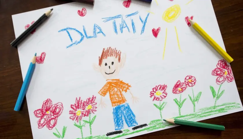Dziecko narysowało laurkę dla taty z uśmiechniętą buzią, słońcem i kwiatami.