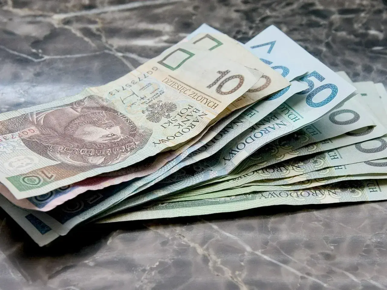 Złotówki na marmurowym blacie. Widać banknoty 10, 50 i inne, które mogą pomóc w pokryciu kosztów związanych z zasiłkiem rodzicielskim.
