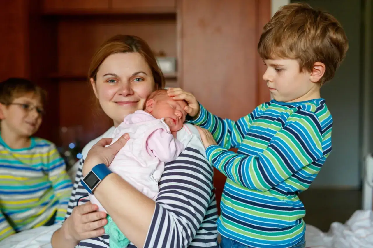 Matka z noworodkiem na rękach, starszy syn delikatnie dotyka główki dziecka, a drugi syn patrzy z boku. Widać ambiwalencję uczuć starszych dzieci.